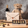 This gift features a bottle of wine, a love letter box of chocolate truffles, a plush teddy bear toy, and a live-edge serving board for presentation.